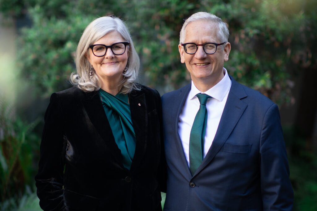 The Governor-General of Australia stands the left side of this photo and her husband on the right, both are smiling for the camera. They stand in front of some green lush background of trees and bushes. They both have grey hair and both wearing glasses. They are wearing dark colours of blue and black and dark green. He wears a white shirt under his suit jacket, and she has a gold pin on her right side of her jacket and pretty earrings